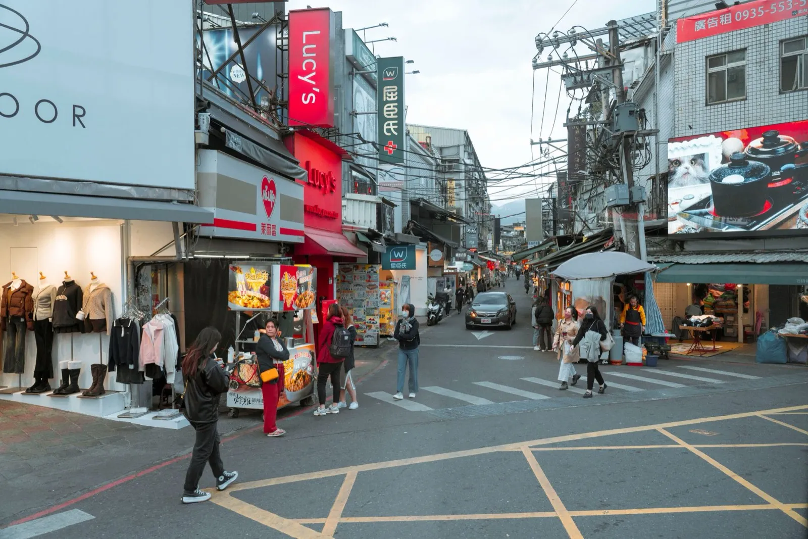 台灣街區路口的服飾店門口掛著外套與上衣,行人走過斑馬線,兩旁招牌林立,天津路服飾商圈逛街